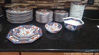 Photo showing stacks of hexagonal shaped dinner plates, salad plates, bread plates, multiple small bowls, and one large serving bowl, all with colorful oriental floral pattern mainly in orange and blue.