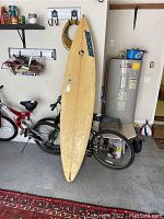 Full view of the yellow fiberglass surfboard leaning against a bike and wall with garage items visible.