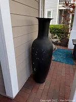 Tall black metal vase standing near a wall outside a house, showing the full item from base to rim.