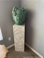 Marble pedestal with the ceramic pot on top placed in a corner near the electrical outlet and window