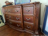 Front view showing curved drawer fronts, nine drawers total (3 large in center and 3 smaller on each side) with pewter-tone handles, ornate carved accents on top and corners.