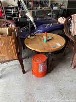 Full view of wood and metal round table showing wood top with scratches and surrounding items including chairs and various garage items.