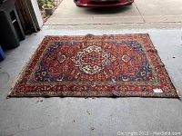 Full view of rectangular Persian-style rug laid flat on garage floor. Predominantly red background with central cream-colored medallion and blue/red geometric and floral border patterns.