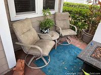 Two outdoor beige cushioned rocking chairs arranged facing each other with a small round table between them on a covered outdoor patio.