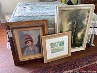 Three framed art pieces leaning against a bed: a portrait oil painting of a girl, a landscape oil painting, and a smaller framed print