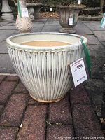 Large cream-colored ceramic planter sitting on paved ground near other planters. Shows vertical ribbed texture and signs of weathering.