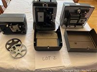 Three vintage movie projectors with their open covers, showing control panels and mechanisms, along with two metal reels on a table.