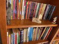 Shelf with around 30 children's books mostly paperbacks in a wooden bookshelf, alongside three figurines including a soldier, sphinx lion, and a girl with watering can.