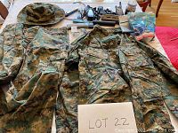 Full view of camouflage outfit with shirt, pants, and hat laid out on table with BB gun supplies in background.