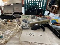 Photo of assorted used tech and stationery items including cords, adapters, pens, mouse, stapler, batteries, and screen protectors laid out on a table.