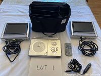 Top-down view showing main DVD player unit, two widescreen LCD displays with power cords, remote control, three cords, two straps, and black carrying case.