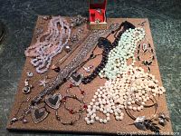 Wide view of assorted costume jewelry including various beaded necklaces, rings, brooch, and bracelets displayed on cork mat.