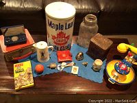 Wide view showing furniture surface holding a Maple Leaf Chips tin can, various small jars, tins, toys, a Mad Magazine booklet, and a commemorative coin case.