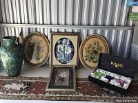 Photo showing a large greenish-blue 3-D floral vase-shaped decor, three framed floral artworks (two oval dried flowers and one square 3-D white flowers), and a black Bombay box on a floral patterned runner.