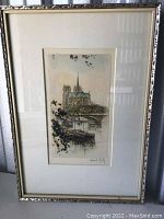 Photo of framed artwork showing full view with silver decorative frame and matting. Image depicts Notre Dame Cathedral and riverboat.