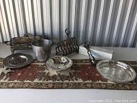 Wide shot showing all the silver plated items on a floral patterned cloth including trays, chafing dish, ice bucket, toast rack, covered dish, and plate.