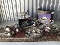 Photo showing six silver plated items arranged including gravy boat on tray, pitcher, covered serving dish, sugar bowl and creamer, and round tray.