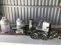 Photo showing various small kitchen appliances including two Cuisinart food processors, Braun electric can opener, and multiple metal discs and accessories for food processing.