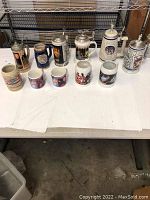 Eleven steins and mugs displayed on a white cloth on a table showing a variety of themes including Star Wars, Star Trek, Budweiser, and German styles.