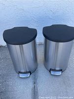 Two stainless steel kitchen garbage cans outside against wall, both showing black lids and foot pedals
