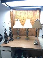 Five table lamps on desk against wood panel wall; two dark ribbed metal lamps without shades, two ornate antique-style lamps with cream shades and beads, one smaller bronze-tone lamp with square beige shade.