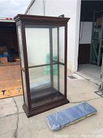 Front view of the tall display cabinet showing the dark wood frame, glass panels, and crown molding at the top. Glass shelves visible inside through the glass door.