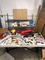 Overview of the large lot with variety of diecast and plastic cars, trucks, trailers, and M&M figures on table and shelf.