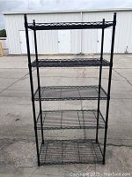 Front view of black metal rack shelf showing five wire shelves and vertical supports outdoors on paved surface.