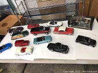 Photo showing 10 diecast cars on a white table and one boxed model in black packaging. Cars vary in color and style including classic and sporty models.