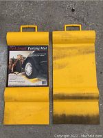 Two yellow Park Smart parking mats showing handles and slotted curved design to hold tires in place, one has packaging label intact