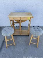 Full view of wooden gate leg folding leaf table with two drawers and lower shelf, flanked by two wooden stools with tufted gray cushions.