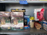 Shelf with two boxes of Malibu Canterbury Collection low voltage mission style path lights, heavy duty adhesive tube, Schlage keypad deadbolt, rolls of masking tape and duct tape, wooden shims, Delta towel ring in package, and magic erasers.