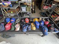 23 assorted hats including sports team hats, promotional hats and various caps displayed on a black table in a garage setting.