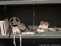 Overall set showing Nintendo Wii console, two Wii remotes, motion sensor bar, steering wheel attachment, cords, and power supply on shelving.
