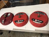 Three red round pressure washer surface cleaners on a table, showing tops with fittings and brand labels