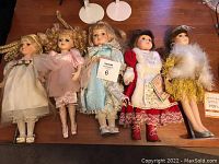 Five porcelain dolls dressed in vintage-style clothing including one in white, pink, blue, red, and yellow dresses, standing on a wooden surface with four white doll stands behind them.