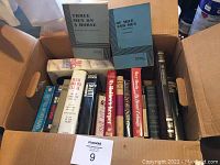 Box of assorted hardcover and paperback books arranged vertically showing the spines with titles visible