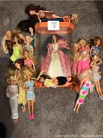 Photo showing sixteen Barbie dolls arranged around an orange-framed clear plastic display case with a Holiday Barbie doll inside. Dolls are dressed in varied casual and formal styles including bright and glittery dresses.