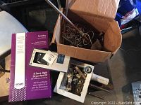 Photo of the lot showing the boxed 8' semi-rigid dryer vent kit, toilet paper holder in box, and open box of mixed plumbing fixtures and parts.