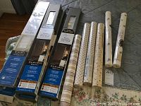 Photo showing five unopened boxes of vinyl blinds, three identified as room darkening, and six unopened rolls of wallpaper with floral patterns on a bedspread.