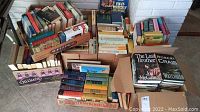 Several boxes and crates filled with books of various genres including fiction and non-fiction, showing colors and sizes of spines, some stacked vertically and horizontally.