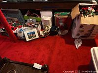 Overall view of Christmas and holiday decor items stored on red carpeted floor including boxes, bags and various decorations