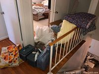 Overview of the comforters stacked on wood railing, blue luggage and black iron on the floor near a door, with some textiles and cardboard boxes in background.