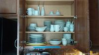 Kitchen cabinet with shelves containing neatly stacked dishes, cups, saucers, coffee pots, and serving platters.