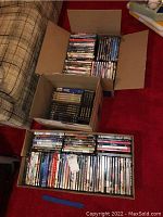 Three open boxes filled with various DVD movies arranged side by side on red carpet near couch.