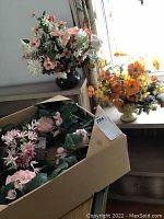 Three floral arrangements, including one in a black vase, one yellow and orange flower arrangement in a yellow matte bowl, and a pink faux flower wreath in a box