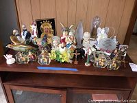 Overview of the lot showing numerous figurines including religious figures, birds, and decorative plaques on a brown wooden surface in front of a wood-paneled wall.