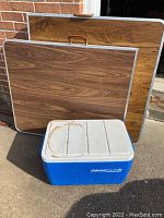 Two folding tables and a blue Coleman PolyLite 34 cooler placed outdoors on pavement. Tables have wood grain laminate tops and aluminum edges.