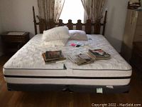 Full view of king size bed with mattress on box springs with wooden headboard and pillows plus assorted magazines on top.