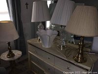 Photo showing four lamps on a dresser with a white scalloped top side table visible underneath; lamps have brass and clear glass bases with white lampshades.